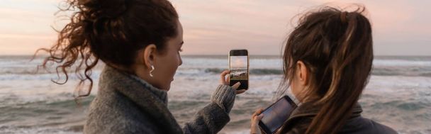 Amigos tirando fotos em smartphones perto do mar durante o pôr do sol, banner  - Foto, Imagem