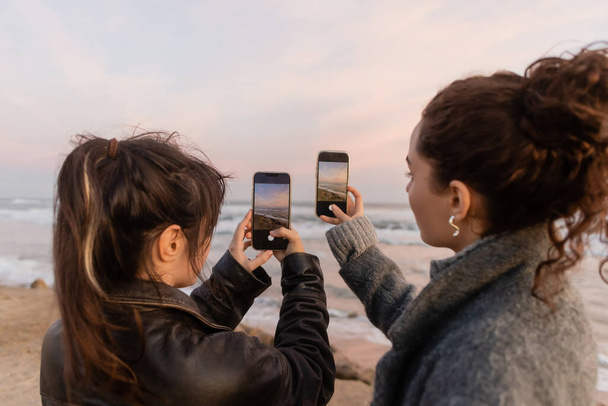 Young friends taking photo on smartphones while standing on beach in Spain  - Foto, Bild