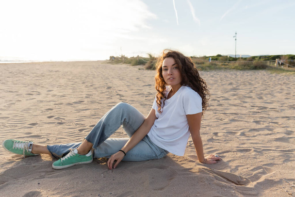 Цікава жінка в джинсах і футболці сидить на пляжі в Іспанії.  - Фото, зображення