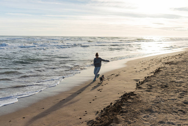 Giovane donna che corre vicino al cane carlino sulla spiaggia sabbiosa di Barcellona  - Foto, immagini