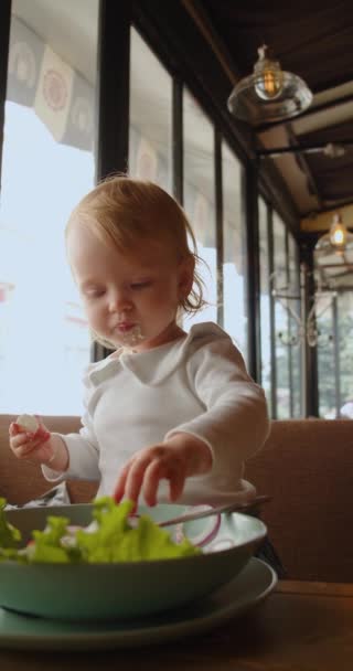 Маленькая голубоглазый девочка ест - она берет еду с тарелки с руками и мажет ее на лице. Отснятый в 4k по вертикали. - Кадры, видео