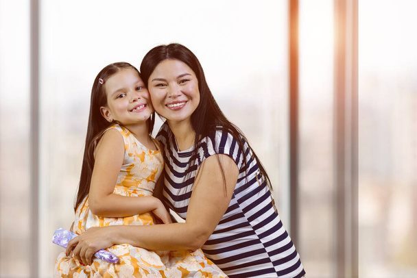 Portrait of a happy asian woman with her adopted caucasian little daughter. Cute little girl with her mother indoors. Blurred windows background. - Foto, immagini