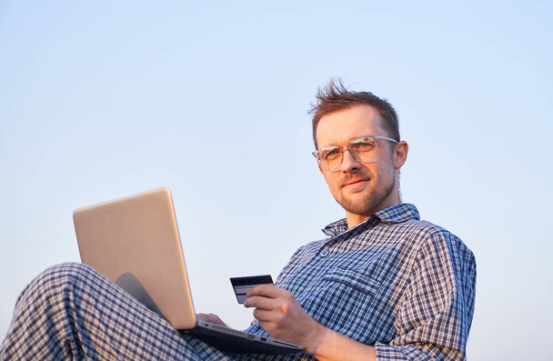 Online shopping concept: caucasian bearded male in eyeglasses making shopping outdoor using laptop and credit card wearing sleep wear. Clear blue sky at background. High quality image - Foto, Imagem