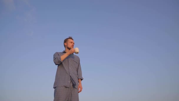 Adorable caucasian male standing outdoor in nightwear looking forward, drinking coffee or tea. Clear blue sky at background. High quality 4k video footage - Imágenes, Vídeo