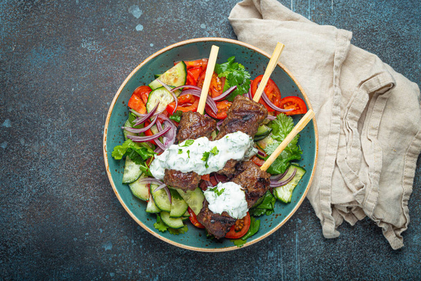 Grilled skewer meat beef kebabs on sticks served with fresh vegetables salad on plate on rustic concrete background from above. Traditional Middle Eastern and Turkish dish Kebab, space for text - Foto, Imagen