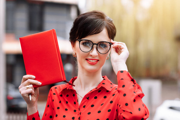 Σίγουρη χαμογελαστή γυναίκα σε γυαλιά δείχνει κόκκινο σημειωματάριο χαρτί με mock up και κοιτάζοντας κάμερα σε εξωτερικούς χώρους στο δρόμο φόντο. Γυναίκα σε κόκκινο πουκαμίσα. Έννοια της διαφήμισης - Φωτογραφία, εικόνα
