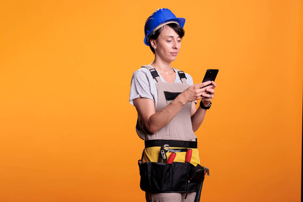 Distracted female builder using smartphone app in studio, scrolling through social media online website. Woman construction worker browsing internet network, wearing helmet and uniform. - Photo, Image