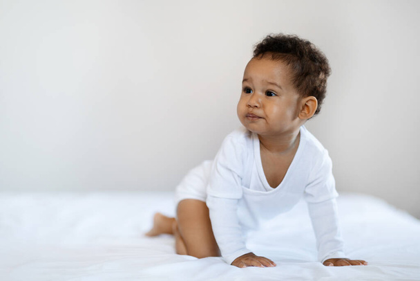 育児。かわいい黒の赤ちゃん男の子か女の子は家でベッドの上でクロールし、離れて見て,ベッドルームで休息を身に着けている愛らしい小さなアフリカ系アメリカ人幼児の肖像画,スペースをコピー - 写真・画像
