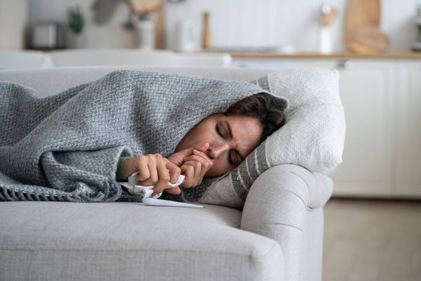 Хвора виснажена жінка лежить на дивані, загорнутому в ковдру і кашляє після зараження небезпечним грипом або вірусною інфекцією. Страждання нещасної дівчини з хусткою і термометром потребує допомоги лікаря - Фото, зображення