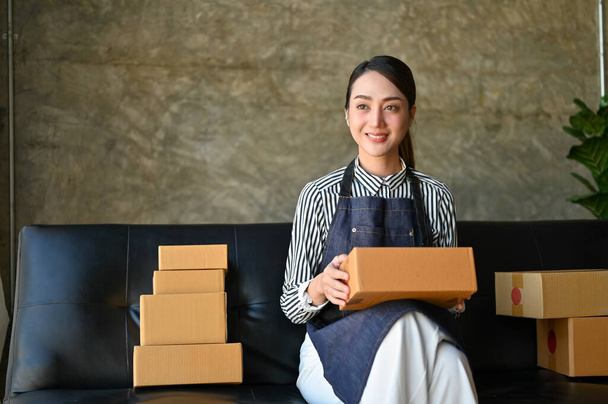 Attractive millennial Asian female e-commerce business owner or online store owner sits on sofa with her shipping packages. - Fotó, kép