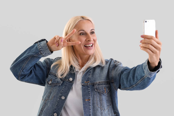 Mature blonde woman with mobile phone taking selfie on light background - Photo, Image