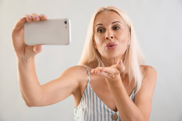 Mature blonde woman with mobile phone taking selfie on light background - Foto, afbeelding