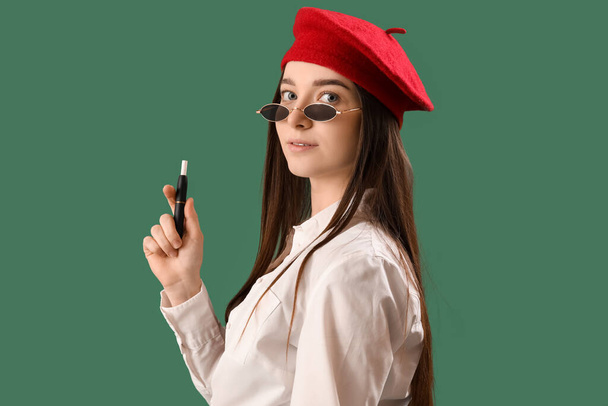 Стильна молода жінка з одноразовою електронною сигаретою на зеленому фоні, крупним планом
 - Фото, зображення