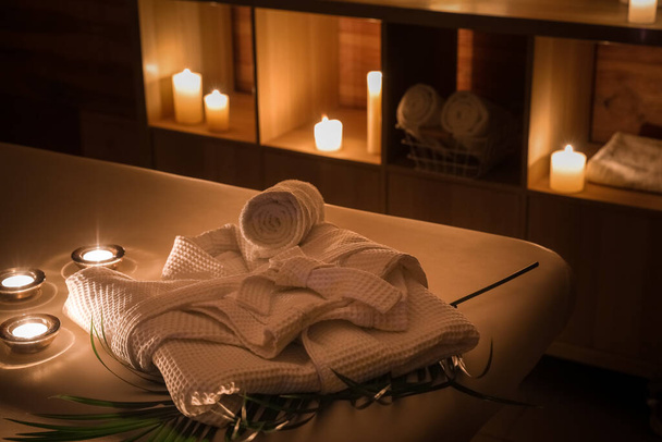 Bathrobe with palm leaf and burning candles on couch in dark spa salon, closeup - Foto, imagen