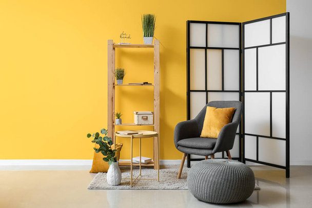 Интерьер комнаты с раскладным экраном, креслом и пуфом возле желтой стены - Фото, изображение