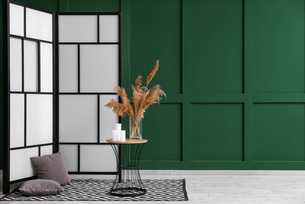 Складной экран, подушки и стол с пампасами трава в вазе возле зеленой стены - Фото, изображение