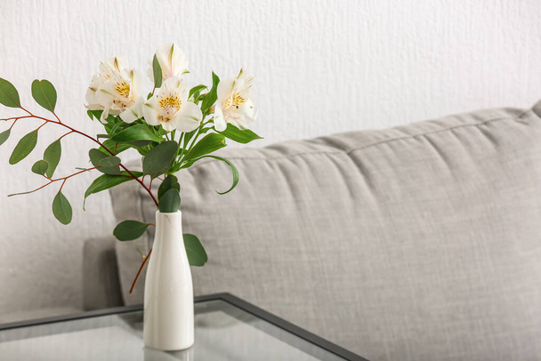 Vase with bouquet of alstroemeria flowers on glass table near grey sofa - Foto, Imagen