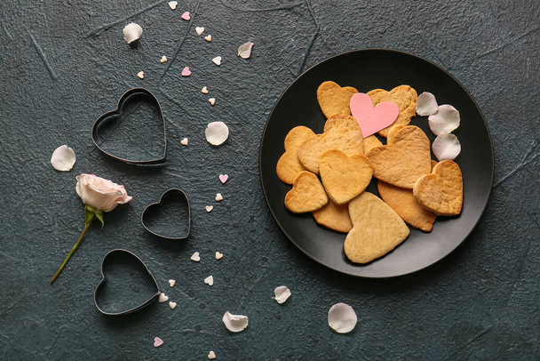 Σύνθεση σαλονιού με πιάτο από νόστιμα μπισκότα σε σχήμα καρδιάς, κόφτες και ροδοπέταλα σε σκούρο χρώμα φόντο. Γιορτή του Αγίου Βαλεντίνου - Φωτογραφία, εικόνα