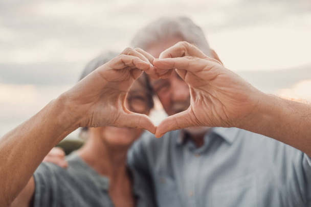 Close up portrait happy sincere middle aged elderly retired family couple making heart gesture with fingers, showing love or demonstrating sincere feelings together outdoors, looking at camera. - Foto, Imagen