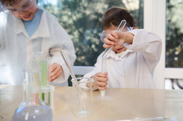 小さな女の子の手に選択的な焦点を当て、化学クラスの間に、学校の研究室で、化学物質を含む他の試験管に試験管から試薬を注ぎます。学校、教育、学習の概念 - 写真・画像