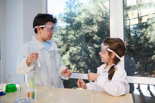 Fascinated teenage boy and primary school girl in safety goggles and lab coats. learning chemistry, conducting experiments, having fun at chemistry class in school scientific laboratory. Education - Zdjęcie, obraz