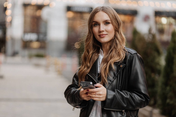 Hermosa mujer de pelo rubio joven sonriente en una chaqueta de cuero negro usando el teléfono y haciendo videollamada en un fondo de la calle de la ciudad - Foto, imagen