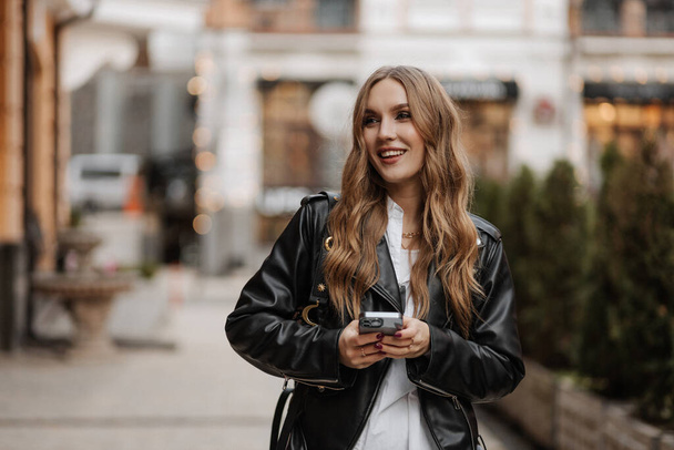 Hermosa mujer de pelo rubio joven sonriente en una chaqueta de cuero negro usando el teléfono y haciendo videollamada en un fondo de la calle de la ciudad - Foto, imagen