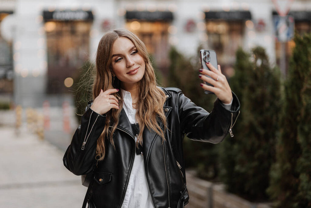 Όμορφη χαμογελαστή νεαρή ξανθιά γυναίκα με μαύρο δερμάτινο μπουφάν χρησιμοποιώντας το τηλέφωνο και κάνοντας βιντεοκλήση σε φόντο του δρόμου της πόλης - Φωτογραφία, εικόνα