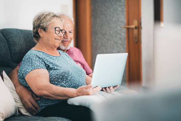 自宅で一緒にラップトップを使用してリラックスしているシニアカップル。成熟した退職者のための新しい現代的なライフスタイル。屋内レジャーでソファの上に座ってコンピュータとインターネット接続を持つ男と女 - 写真・画像