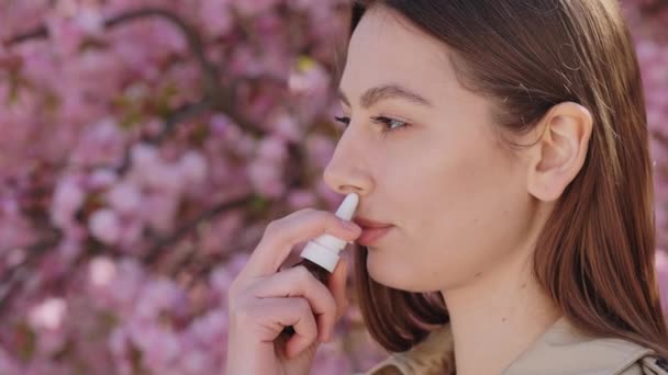 Side view of sick young woman using nasal spray outdoors because of tree pollen allergy. Irritated brunette with health problems during seasonal flowering. - Felvétel, videó
