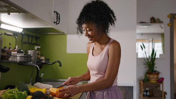 African American woman cuts fresh orange with knife dancing in kitchen. Housewife enjoys making breakfast from organic products - Photo, image