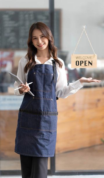 Красивая азиатская молодая бариста в фартуке держит планшет и стоит перед дверью кафе с открытой вывеской. Концепция запуска бизнеса... - Фото, изображение