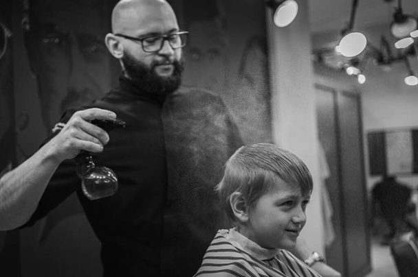 Happy cute fair-haired preschool boy getting a haircut. Children's hairdresser with scissors and comb cuts a little boy's hair in a room with a loft interior - Foto, Bild