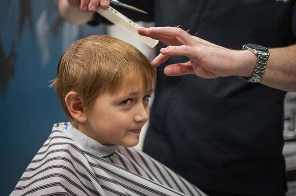 Happy cute fair-haired preschool boy getting a haircut. Children's hairdresser with scissors and comb cuts a little boy's hair in a room with a loft interior - Fotó, kép