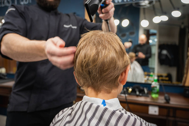 Happy cute fair-haired preschool boy getting a haircut. Children's hairdresser with scissors and comb cuts a little boy's hair in a room with a loft interior - Foto, afbeelding