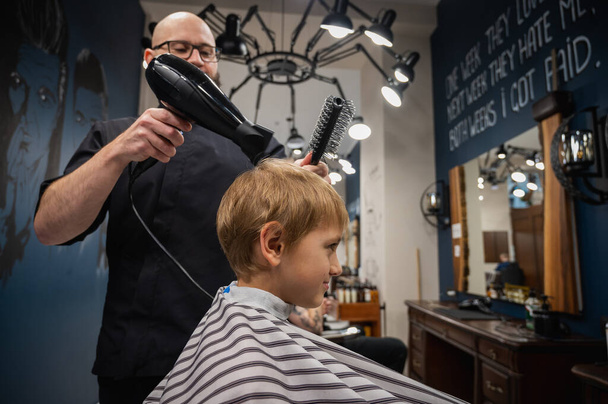 Happy cute fair-haired preschool boy getting a haircut. Children's hairdresser with scissors and comb cuts a little boy's hair in a room with a loft interior - Photo, image