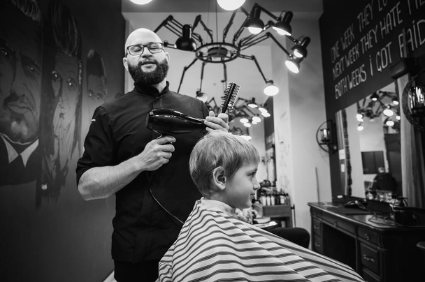 Happy cute fair-haired preschool boy getting a haircut. Children's hairdresser with scissors and comb cuts a little boy's hair in a room with a loft interior - Photo, Image