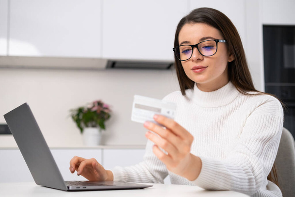 Woman enters card details into laptop to buy educational courses. Brunette lady sits in the kitchen making online payment closeup - Foto, imagen