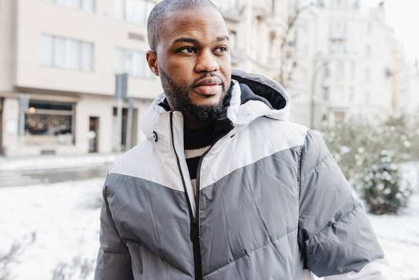 Young African man in casual white and gray clothes, sport jacket. Winter season street portrait in park with snow. Lifestyle, city life concept, copy space - Foto, afbeelding