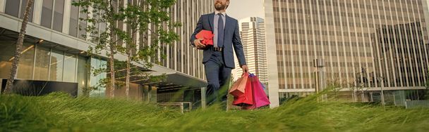 обрезанный бизнесмен в костюме держать пакеты и подарочная коробка ходить за пределами офиса, покупки. - Фото, изображение