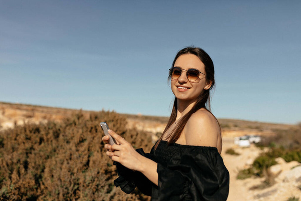 Charming adorable stylish woman with happy smile and dark hair wearing black dress with bare shoulders in sunglasses using smartphone and posing at camera while travelling in mountains  - Fotografie, Obrázek