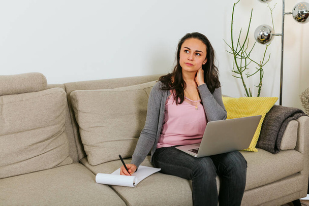 Λατίνα κυρία που κάθεται στον καναπέ δουλεύοντας με φορητό υπολογιστή στα πόδια και σκέφτεται τις σημειώσεις - Φωτογραφία, εικόνα