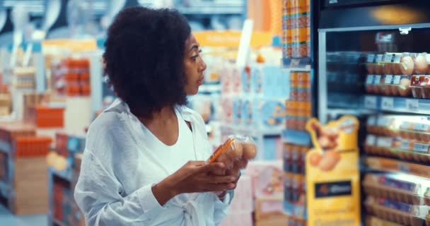 Многонациональная темнокожая женщина, читающая этикетку на упаковке с органическими яйцами животных во время покупки продуктов - Кадры, видео