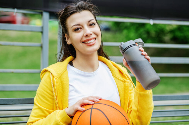 Portrait of young woman sitting on bench with basketball and drinking water during break. Hobby and sport concept. - Valokuva, kuva