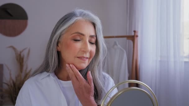Smiling beautiful aging 50s yeas old lady skin care at home moisture looking at mirror reflection touch face cream facial anti-wrinkled cosmetic 60s senior mature woman touching cheeks lifting wrinkle - Footage, Video