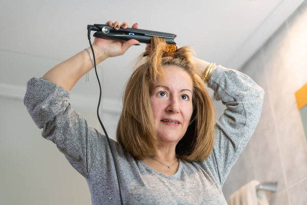 Mature white woman fixing her hair in front of the mirror with a hair straightening device - Foto, afbeelding
