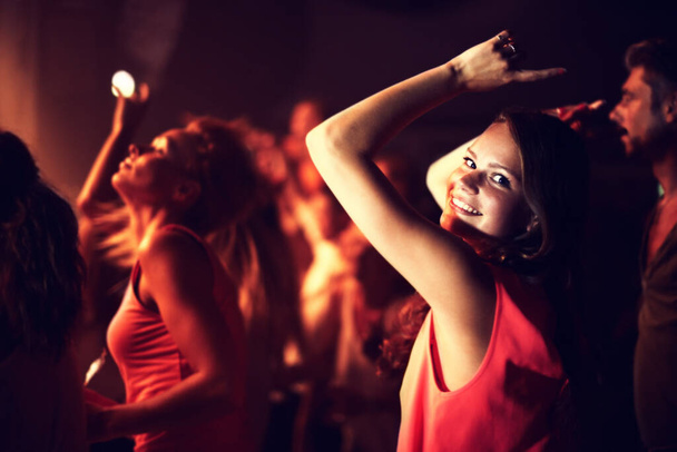 Απολαμβάνω τις δονήσεις στο κλαμπ. μια νεαρή γυναίκα που χορεύει με τα χέρια ψηλά με ανθρώπους στο βάθος - Φωτογραφία, εικόνα