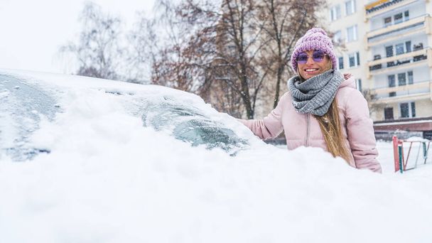 Πορτρέτο μιας νεαρής γυναίκας κοντά στο αυτοκίνητο καλυμμένο με χιόνι. Χειμερινά ρούχα σε ροζ. Υψηλής ποιότητας φωτογραφία - Φωτογραφία, εικόνα