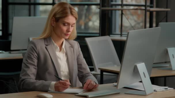 Caucasian middle-aged adult woman mature business female lady businesswoman entrepreneur in office write notes text in notebook on paper typing computer working accounting writing task ideas with pc - Materiał filmowy, wideo