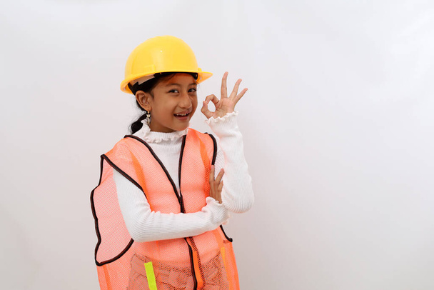 Cheerful Asian little girl in the construction helmet as an engineer standing while showing okay hand gesture. Isolated on white with copyspace - Fotó, kép